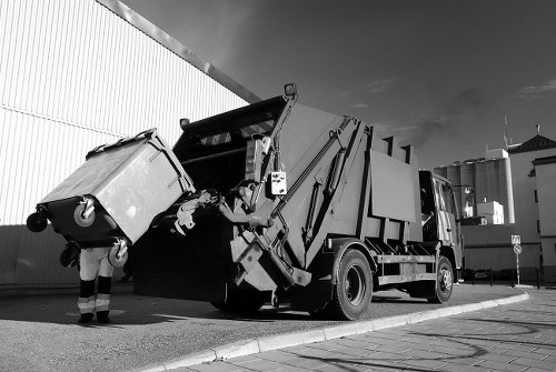 A commercial waste collection vehicle, fitted with a hydraulic lifting arm, is actively engaged in collecting a large, wheeled rubbish bin from a concrete driveway adjacent to a metal-sided industrial or warehouse building. The bin appears to be made of plastic with a textured surface and a flat lid, and is positioned on the left side of the image, being lifted towards the truck's hopper for disposal. The scene is outdoors, with the vehicle parked parallel to the building, under a partly cloudy sky. The vehicle has a streamlined, utilitarian design with visible mechanical components like hydraulic cylinders and hoses, and is primarily painted in dark tones. The surrounding environment includes a curb and paved area, typical of commercial waste collection sites. The image captures the process typical of rubbish removal services within Bounds Green and highlights the practical aspects of waste management undertaken by Commercial Waste Bounds Green, focusing specifically on the collection of commercial rubbish Storage in an urban or industrial setting.