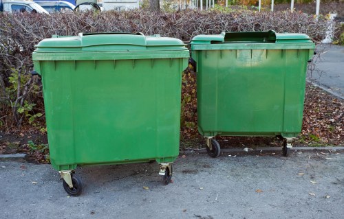 Two large green wheeled rubbish bins made of durable plastic material are positioned side by side on a paved surface. The bin on the left is slightly larger with a flat lid, while the smaller bin on the right has a similar lid and appears to be partially shaded by the larger one. Both bins are set against a background of low, leafless shrubbery and a fence, indicating an outdoor setting, possibly a driveway or service area. The scene suggests a waste collection or disposal context, consistent with commercial waste removal services such as those provided by Commercial Waste Bounds Green, located in the Bounds Green area. The bins' weather-resistant surface reflects natural lighting, with subtle shading revealing its textured finish. The setting appears to be during late autumn or early winter, as evidenced by fallen leaves and the leafless bushes, highlighting the importance of local rubbish management in this area.