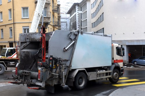 A large commercial rubbish collection van parked on a city street near multi-storey buildings, with a hydraulic lift arm extended towards an upper floor window. The vehicle is predominantly grey with some red accents, and features various hoses, controls, and equipment attached to its side for waste loading. Behind the van, there is a pedestrian crossing with yellow stripes on the road. The environment appears urban, with other parked vehicles visible in the background, including a small car and additional waste-related equipment. The scene suggests active waste collection or removal, typical of professional services provided by Commercial Waste Bounds Green in the Bounds Green area, London, with surrounding buildings and typical street elements visible in the backdrop.
