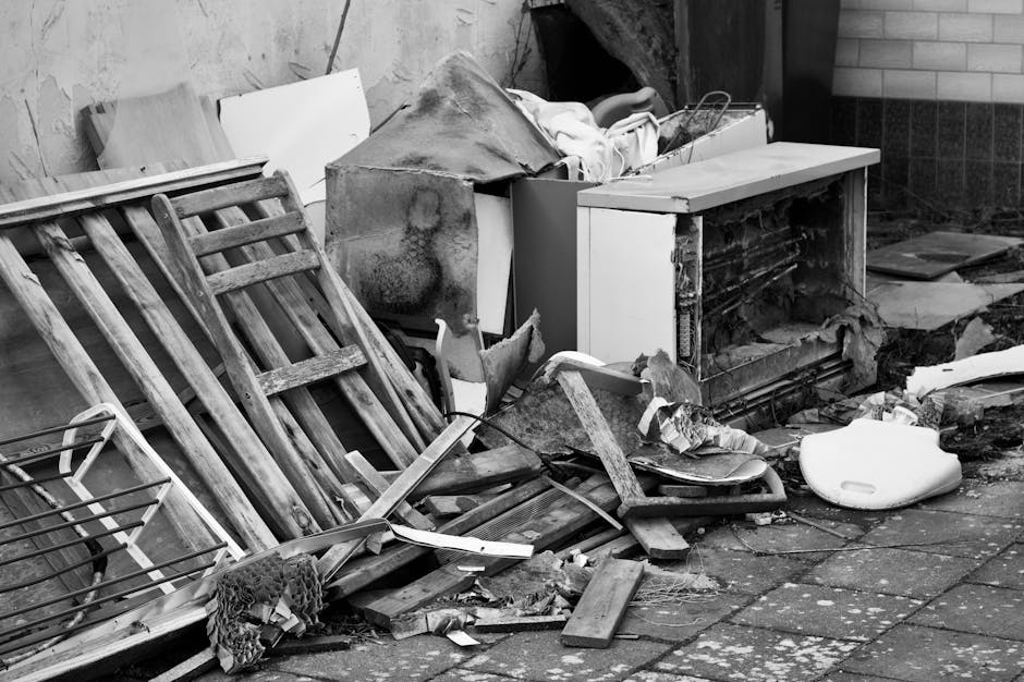 A collection of mixed waste materials and cardboard boxes piled against a rough brick and concrete wall in an outdoor setting, possibly a back alley or garden area. The debris includes flattened and i