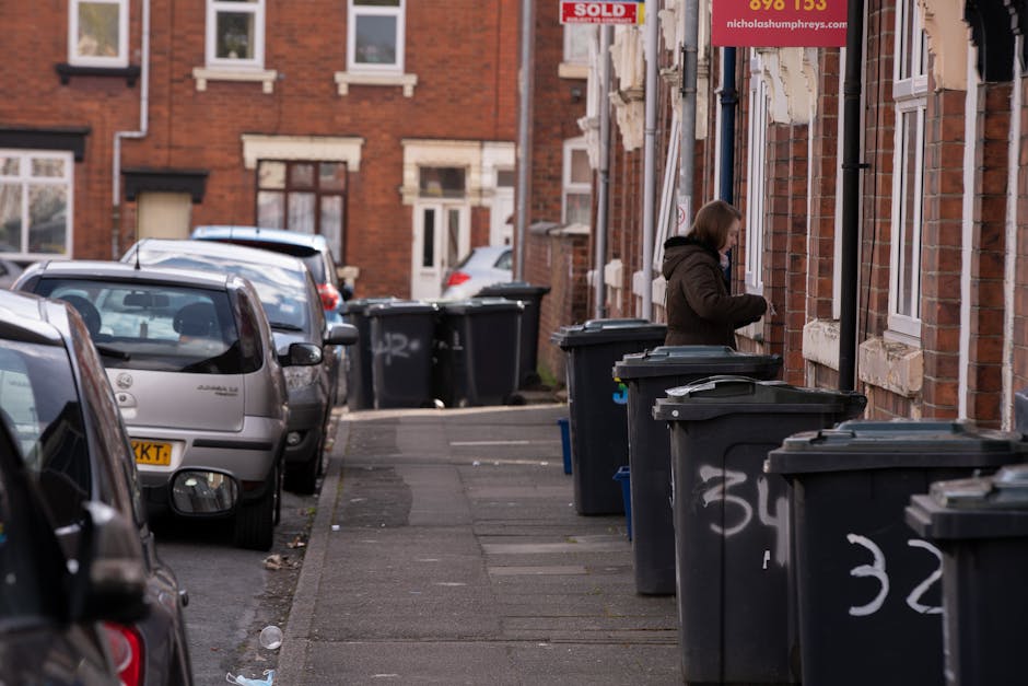 A person disposing of used paper bags into a grey waste bin with a lid, situated on a wooden surface indoors. The paper bags are crumpled and appear to contain food or light refuse. The individual is 