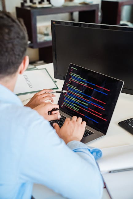 A person wearing a light blue shirt is seated at a desk, working with a laptop that displays lines of colorful computer code on its screen. The individual is holding a pen in their right hand and appe