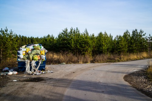 A large blue wheelie bin placed on an unpaved, gravel roadway, filled with assorted household waste including plastic bags, cardboard boxes, and paper, some of which are overflowing and spilling onto the ground around it. The bin appears to be in a rural or semi-rural area, with a backdrop of dense green trees and bushes under a clear, blue sky. The scene suggests improper disposal of rubbish, which may be addressed by professional waste management services such as those offered by Commercial Waste Bounds Green, servicing localities within or near the N11 postcode area, including the surrounding area of London. The gravel road curves gently to the right, with shadows cast by nearby trees, emphasizing the outdoor setting. The overall environment is quiet, with no visible activity, highlighting the importance of proper rubbish removal and sustainable waste management practices to maintain cleanliness and environmental health.