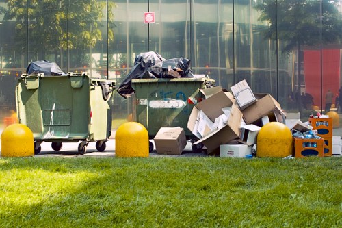 An outdoor scene featuring two large green wheelie bins positioned on a paved area with a reflective glass building in the background. The left bin has a black plastic bag draped over its lid, while the right bin is overflowing with various cardboard boxes, some flattened and others still intact, along with plastic bottles and miscellaneous waste. Surrounding the bins are three yellow plastic barriers that separate the waste area from a well-maintained grass verge. The overall environment suggests a commercial or residential waste collection point, with natural daylight illuminating the scene and highlighting the textures of the discarded materials, primarily cardboard, plastic, and loose bags. This image relates to rubbish removal services provided by Commercial Waste Bounds Green in the local area, exemplifying typical waste storage and disposal points for businesses or nearby properties in London postcode regions.