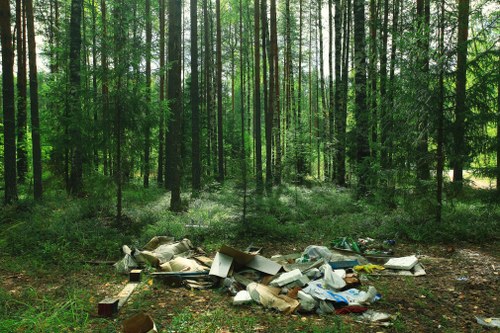 A forest scene with tall, slender trees featuring green foliage and straight trunks, extending into the background where sunlight filters through the canopy. In the foreground, there is a noticeable pile of mixed waste materials including cardboard boxes, plastic containers, paper, and other debris scattered across the ground, which is covered with soil and leaf litter. The waste appears to be dumped illegally on the edge of a wooded area, contrasting with the natural green surroundings. This scene is indicative of unmanaged rubbish disposal near a woodland environment, highlighting the need for professional waste management services such as those provided by Commercial Waste Bounds Green to ensure proper rubbish removal and site cleanliness in the local area near London postcodes, possibly around Bounds Green.